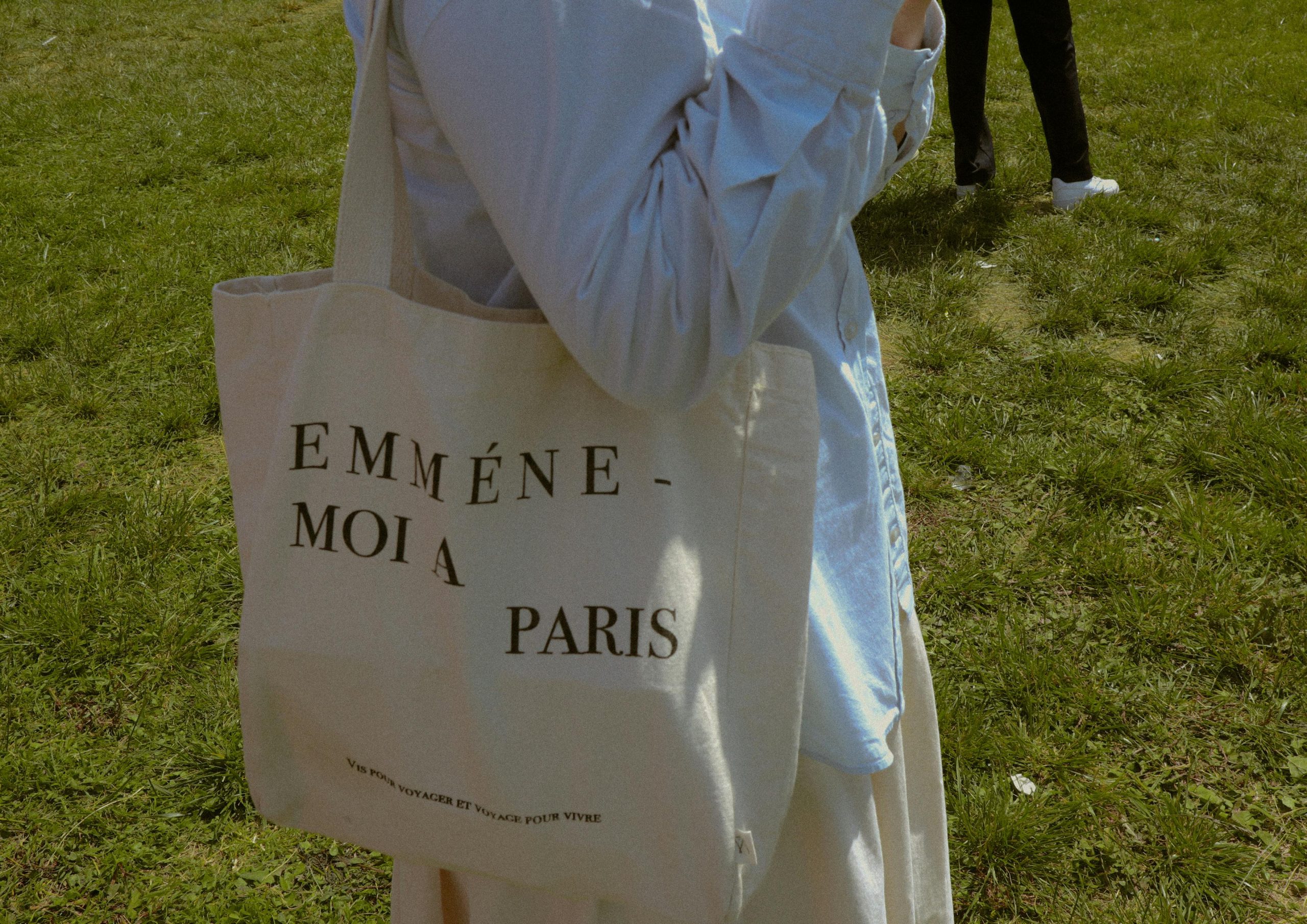 Person carrying a fashion-forward tote bag with French text 'Emmene-moi a Paris' in a grassy outdoor setting.